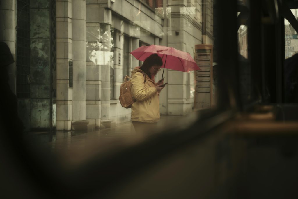 Não encontrou a vaga que deseja? 2 A woman with a pink umbrella checks her phone on a rainy city street, evoking a moody, candid feel.