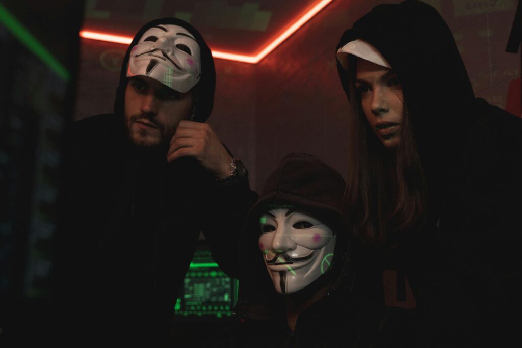 Golpes de emprego (Falsas vagas) 5 A group of people wearing Guy Fawkes masks in a dark tech-themed room, suggesting anonymity and hacking activities.